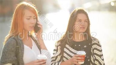 两个可爱的女孩在城里的街道上。 女孩们拿着纸杯喝咖啡。 一个红头发的女孩